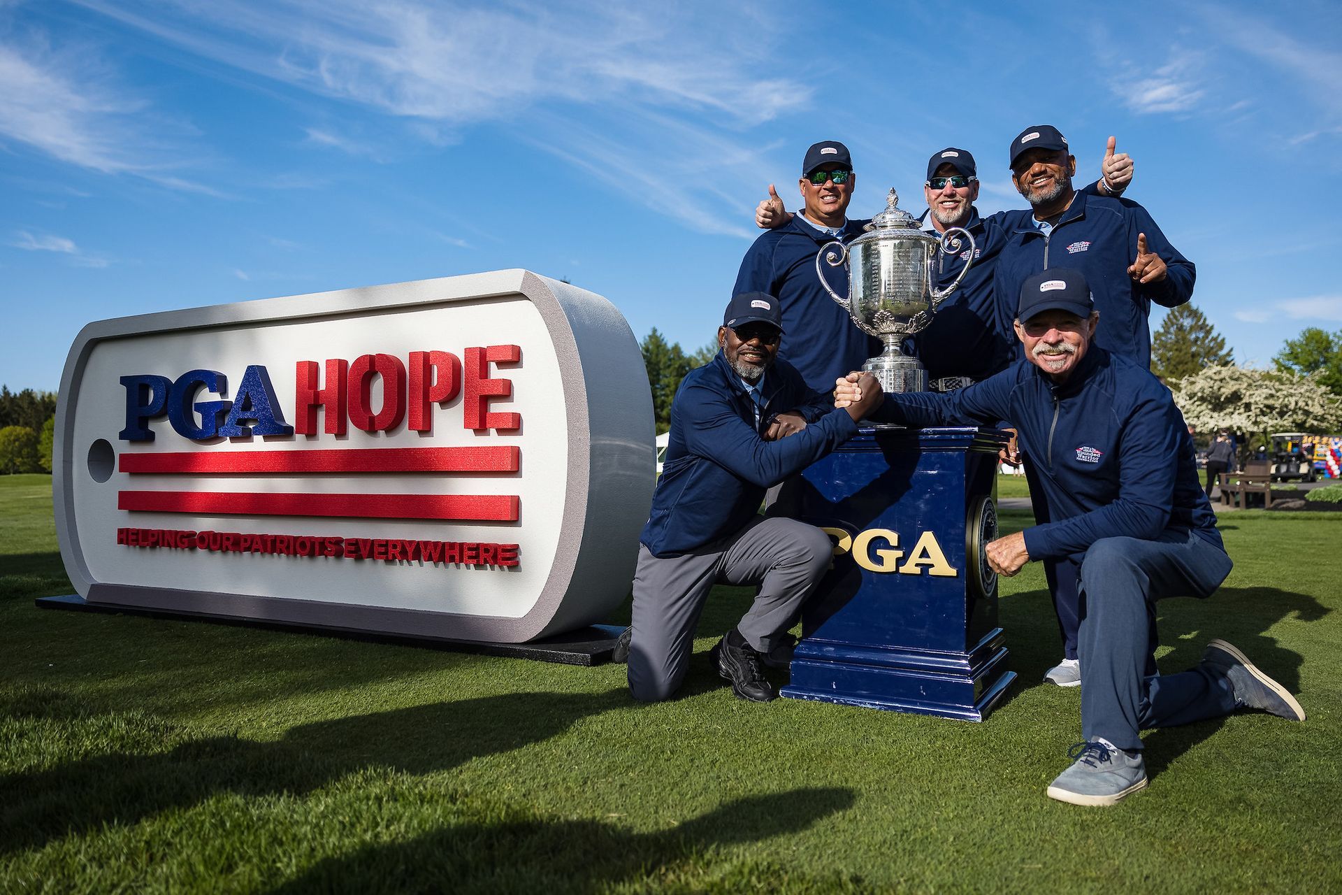 Golf team posing with a trophy beside a large PGA HOPE sign on a grassy field under a blue sky