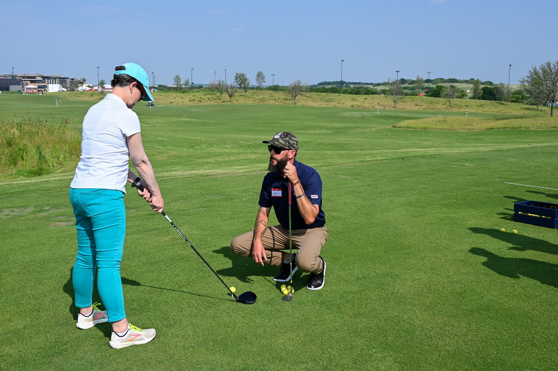 Two golfers on a green fairway, one standing with a club and one kneeling near a ball, under a clear blue sky.