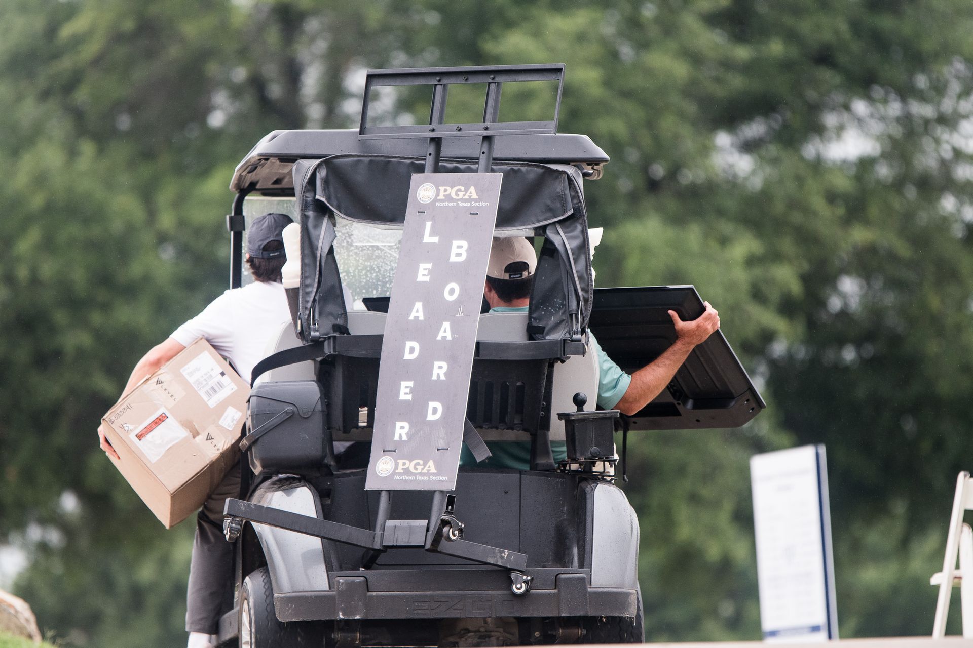 Rear view of a person on a mobility scooter carrying boxes, with a vertical “BOARD” sign, outdoors