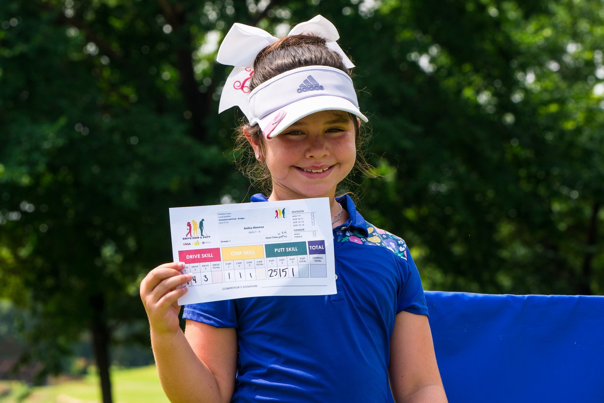 Smiling child in a blue shirt and visor holding a certificate outdoors.