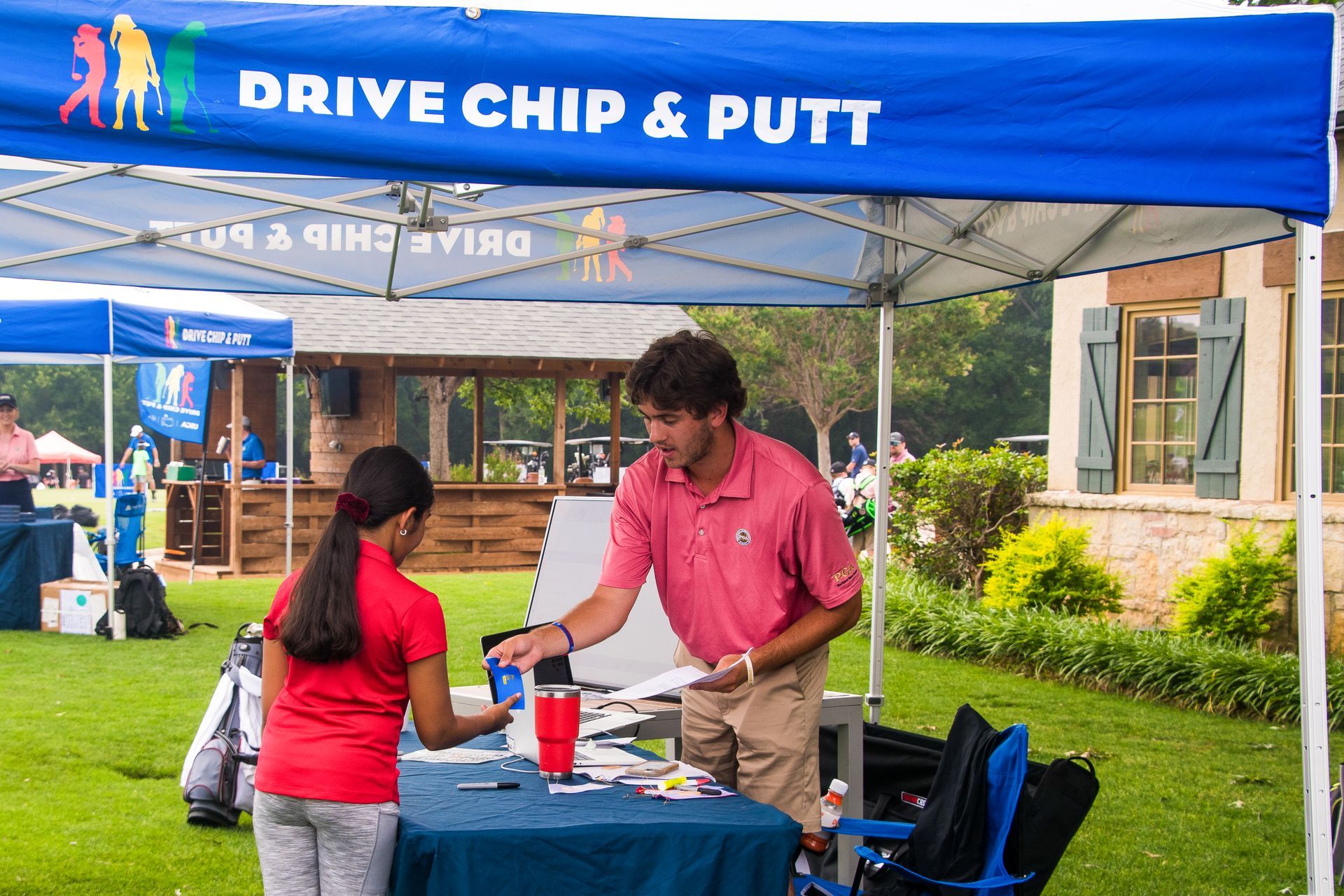 Drive Chip & Putt booth with two people at a table under a blue canopy, outdoors on a rainy day