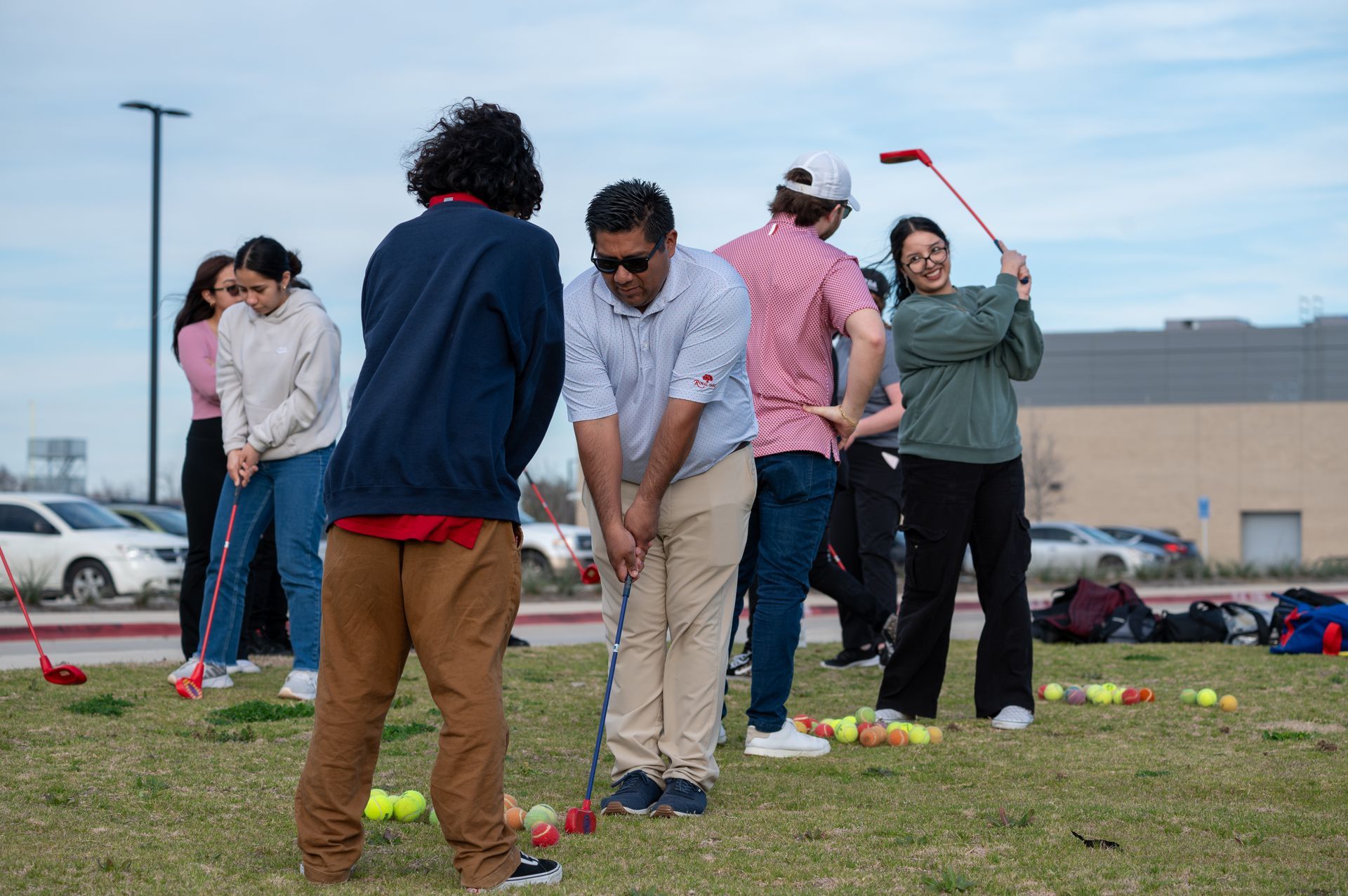 People playing croquet on a grassy field with yellow balls and red wickets under a cloudy sky