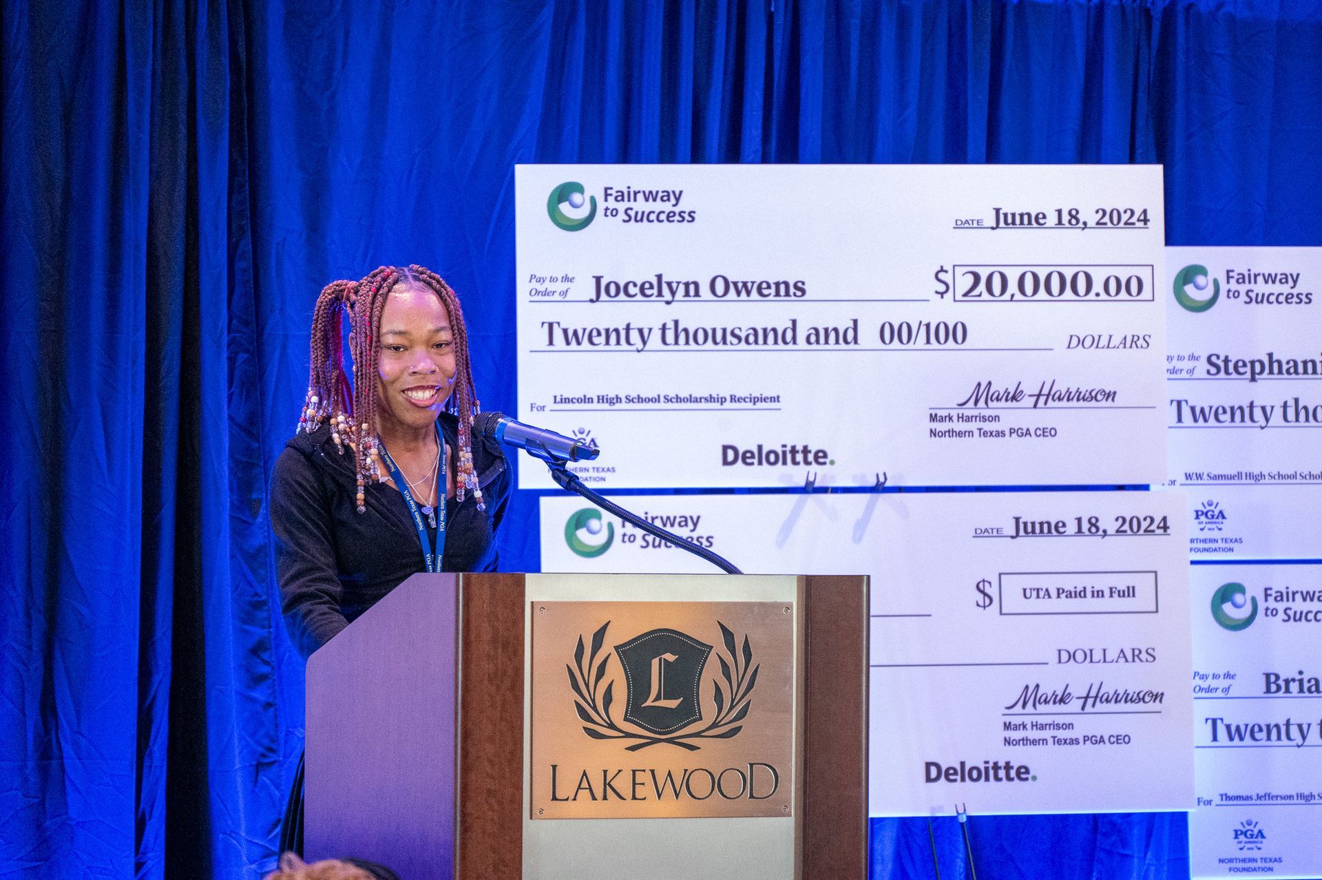 Woman at a podium speaking at a formal event with a blue curtain backdrop and oversized check display