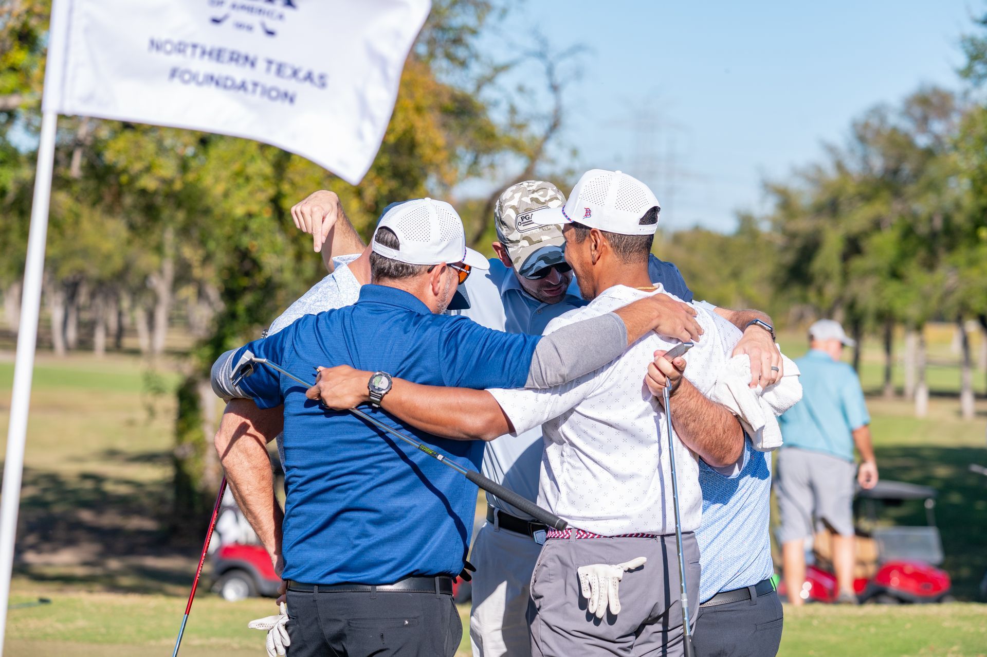Four golfers in white caps and blue shirts huddle on a sunny golf course near a Northern Texas Foundation flag.
