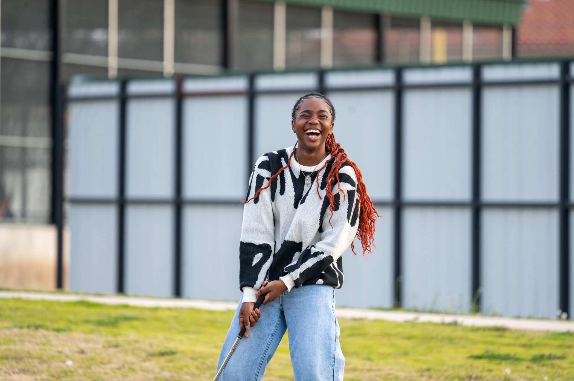Smiling person in a black-and-white sweater and jeans standing outdoors in front of a building