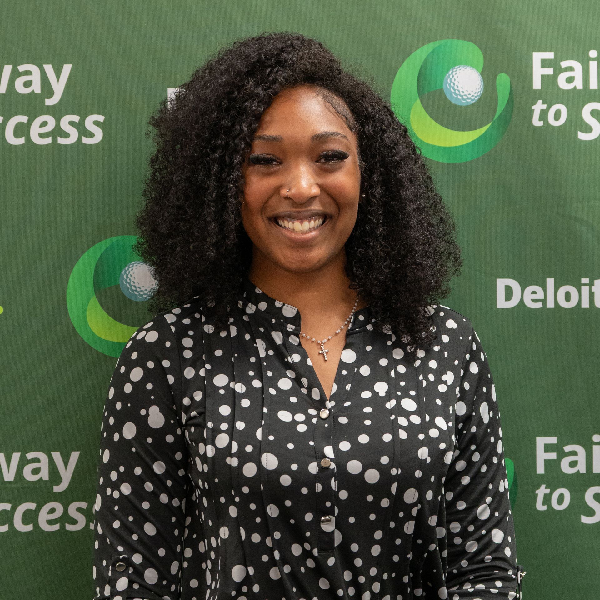 Smiling woman in a black polka-dot blouse standing before a green event backdrop with logos.