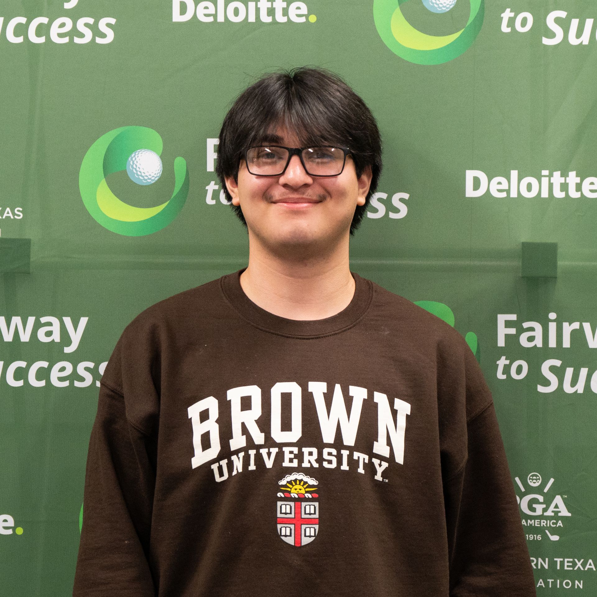 Person in a Brown University sweatshirt standing before a green Deloitte backdrop, smiling.