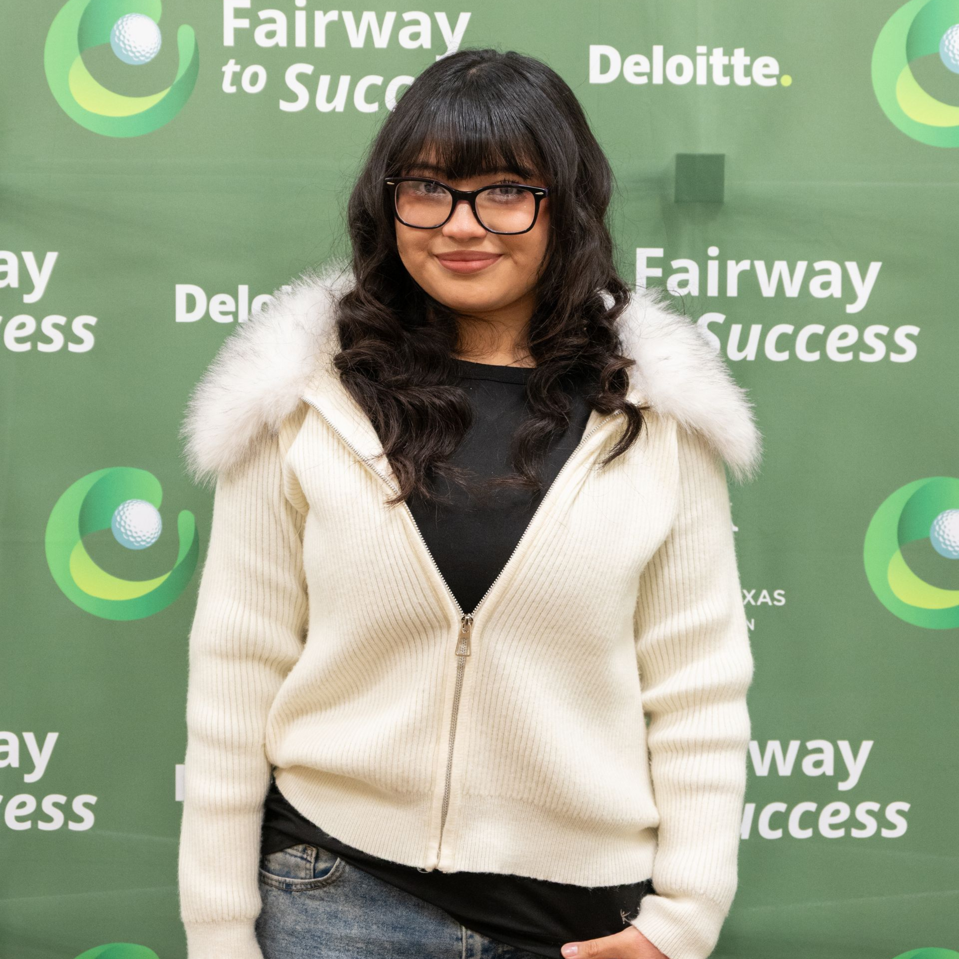 Woman posing at a Fairway to Success event, wearing a cream jacket and glasses, in front of a green Deloitte backdrop