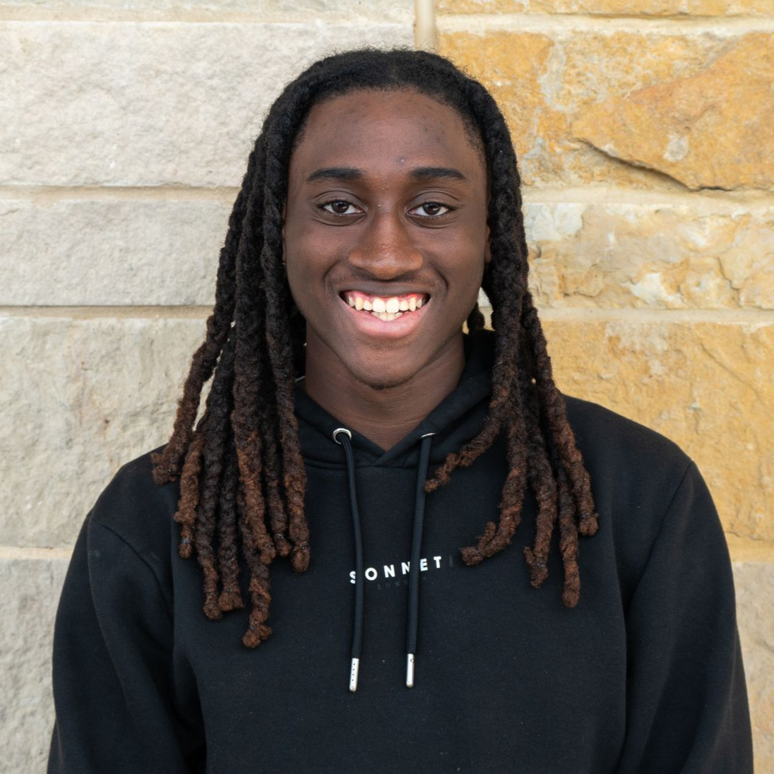 Smiling person with long braided hair wearing a black hoodie, standing against a stone wall.