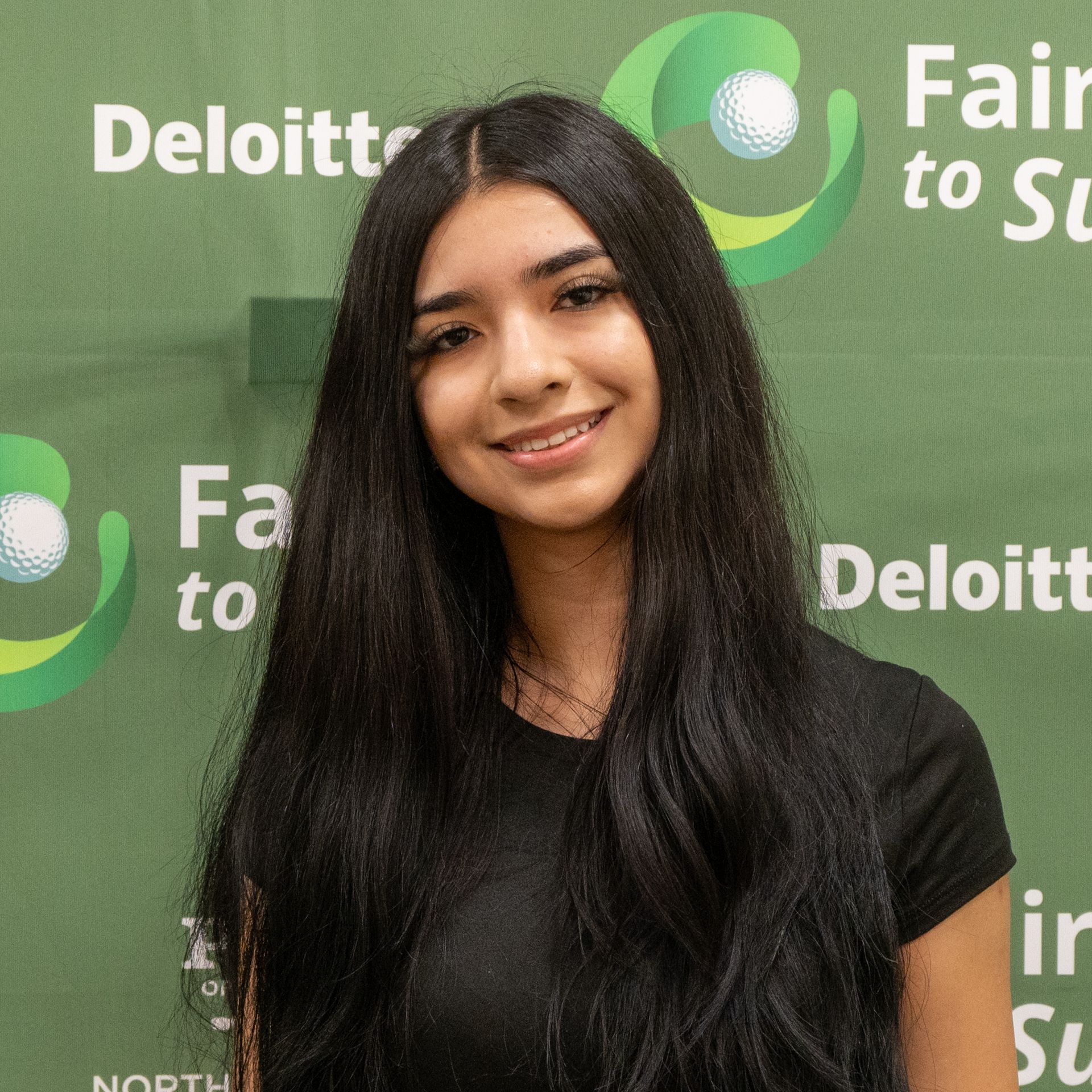 Smiling woman with long dark hair in a black shirt posing before a green Deloitte backdrop