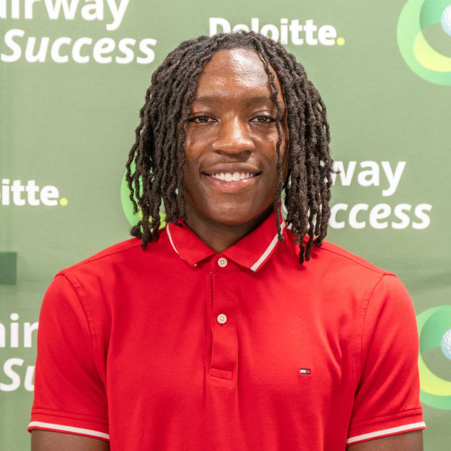 Smiling person in a red polo shirt posing before a green Deloitte “Fairway Success” backdrop