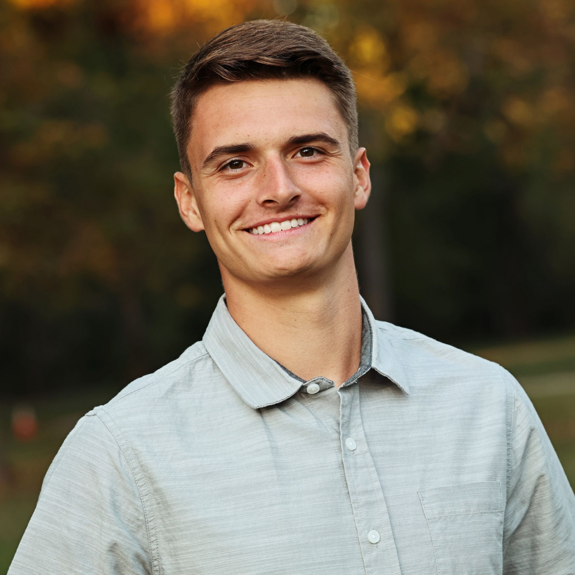 Smiling person in a light gray shirt standing outdoors with a blurred autumn background