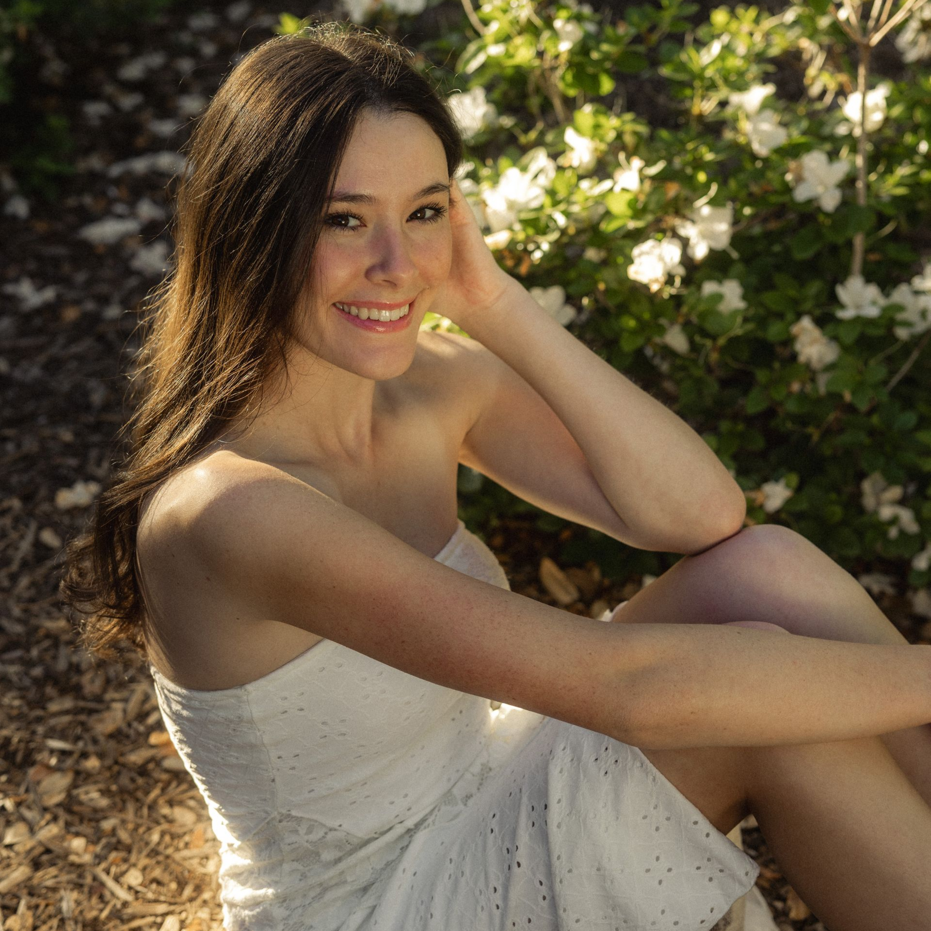 Woman in a white dress sitting outdoors among flowers, smiling at the camera.