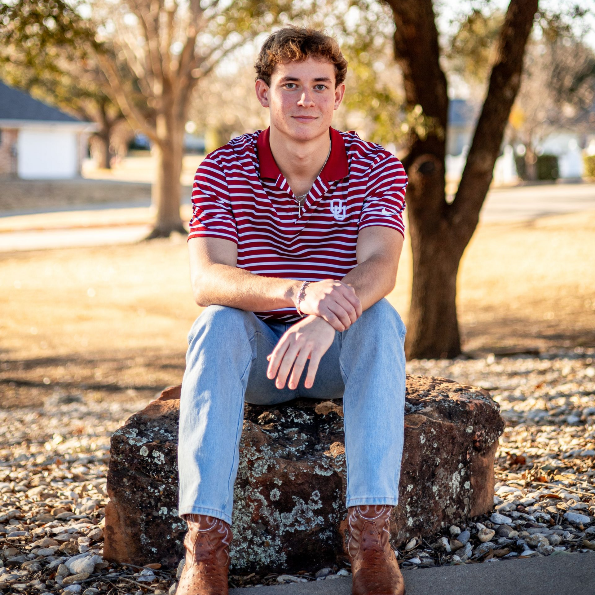 Young man in a red striped shirt sits on a tree stump outdoors in a sunny park