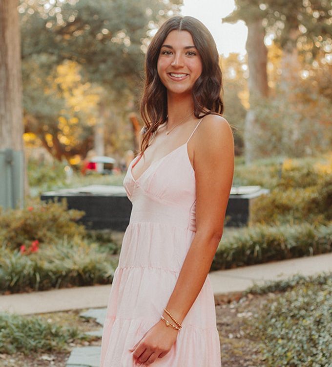 Woman in a light pink dress smiling outdoors in a garden path at sunset