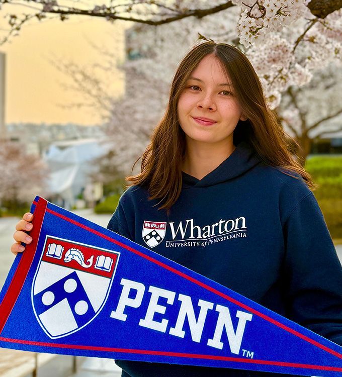 Student in a navy Wharton sweatshirt holding a blue-red PENN pennant outdoors under blooming trees