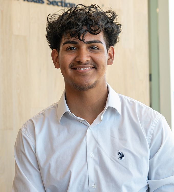 Smiling person in a white shirt standing indoors against a light wood background