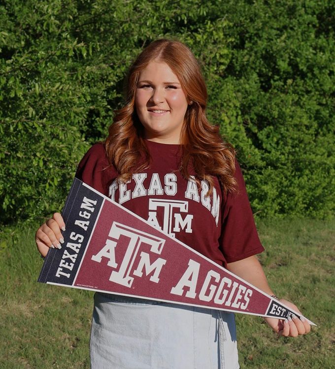 Woman holding a Texas A&M Aggies pennant and sign in front of green trees.