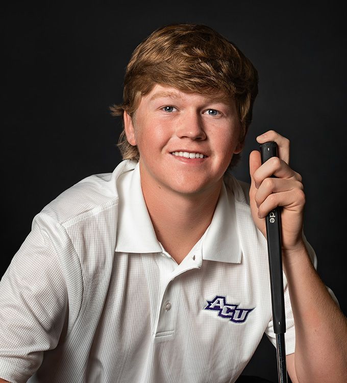 Young athlete in white polo holding a golf club, smiling against a dark studio background