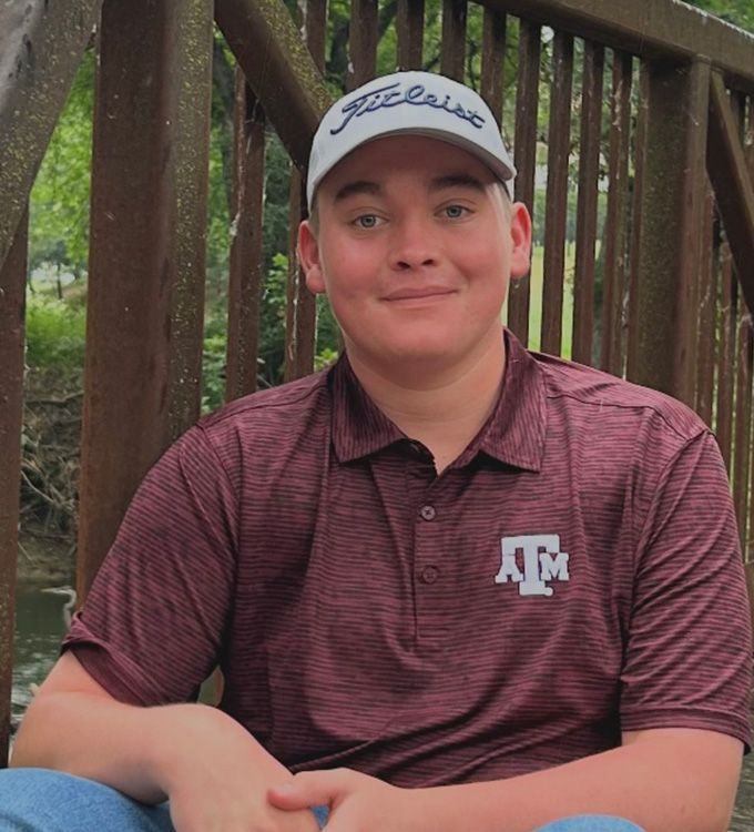 Person in a maroon Texas A&M polo and white cap sitting outdoors on a wooden bridge, smiling.