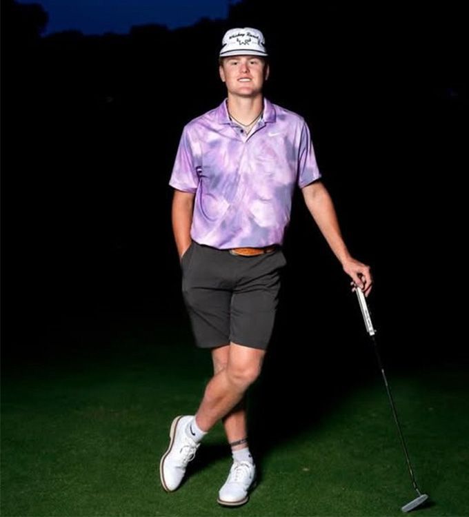 Golfer in a purple shirt and black shorts walking on a dark golf course at night, holding a putter.