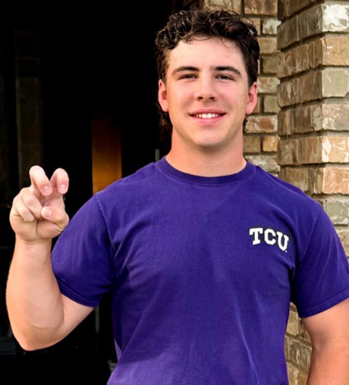 Young person in a purple TCU shirt standing by a brick doorway, smiling and making an air-quote gesture