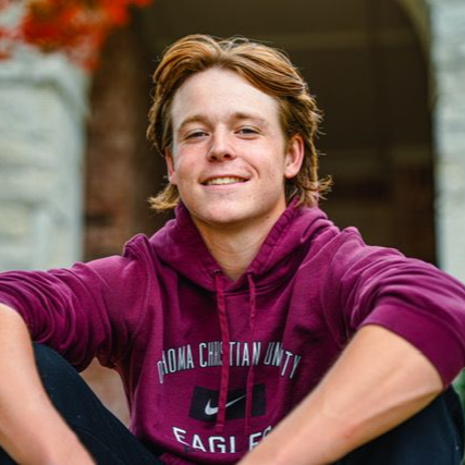 Smiling person in a maroon hoodie sitting outdoors in front of a brick archway