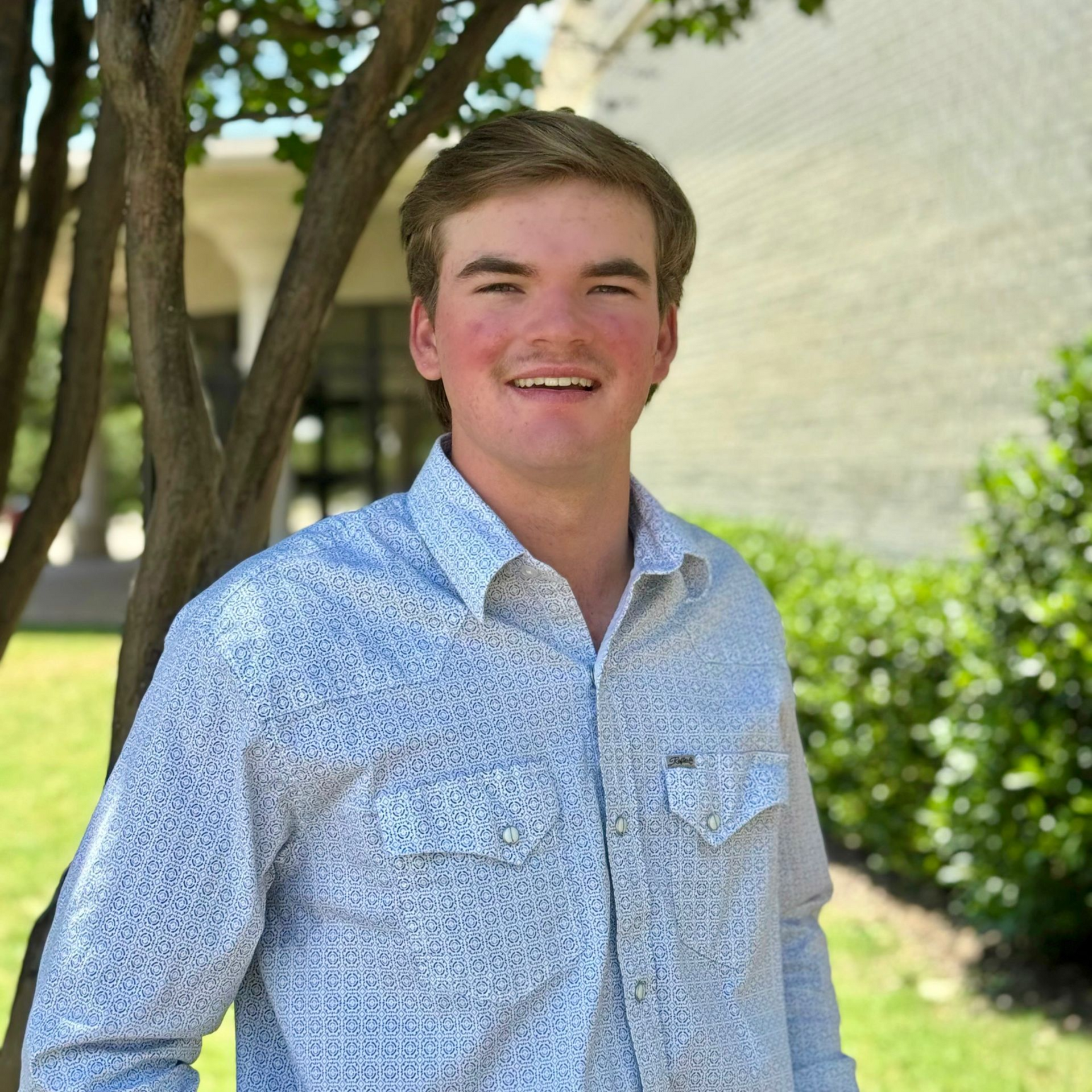 Smiling person in a light blue patterned shirt standing outdoors by a tree and shrubs.