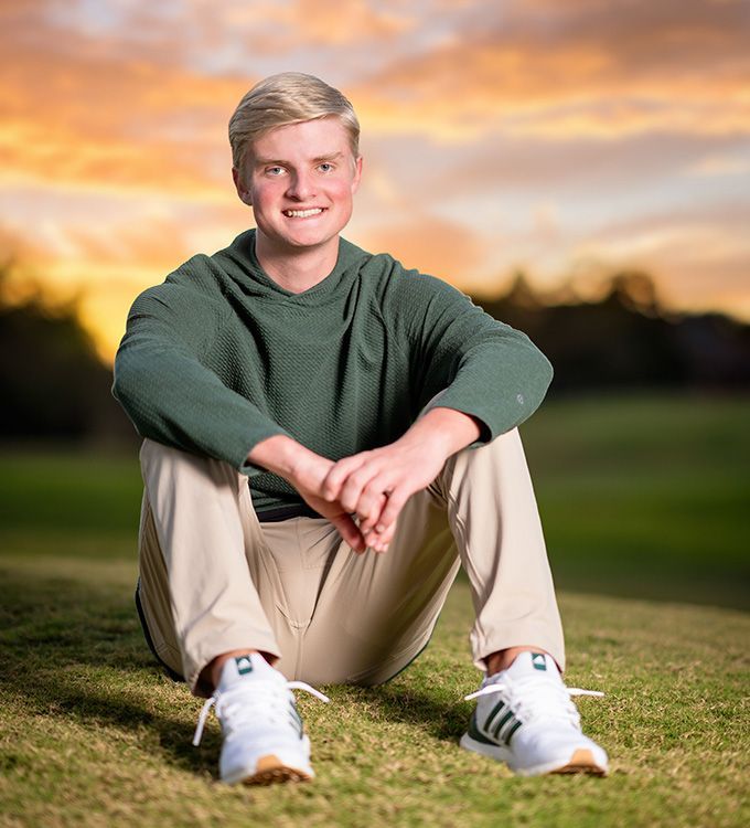 Person sitting on grass at sunset, smiling in a green sweater, tan pants, and white sneakers.