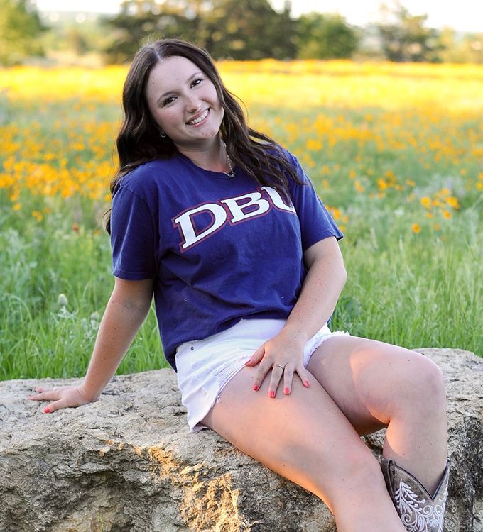 Smiling woman in a purple shirt sitting on a rock in a grassy field with yellow flowers