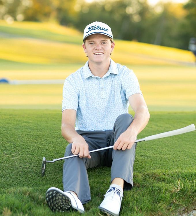 Young golfer sitting on grass holding a putter, wearing a light blue shirt and white cap on a sunny course.
