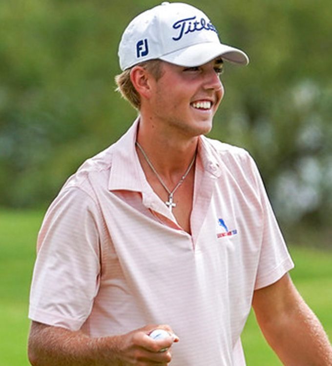 Golfer in a white cap and pink polo smiling on the course, holding a golf ball and club.