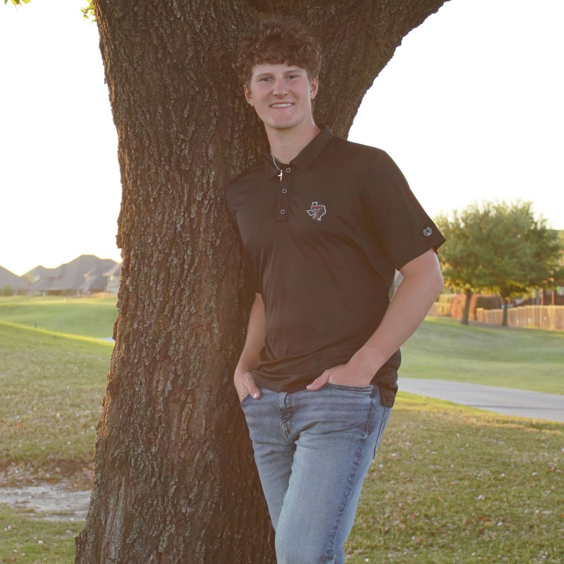 Person leaning against a tree in a grassy park at sunset, wearing a black polo and jeans.