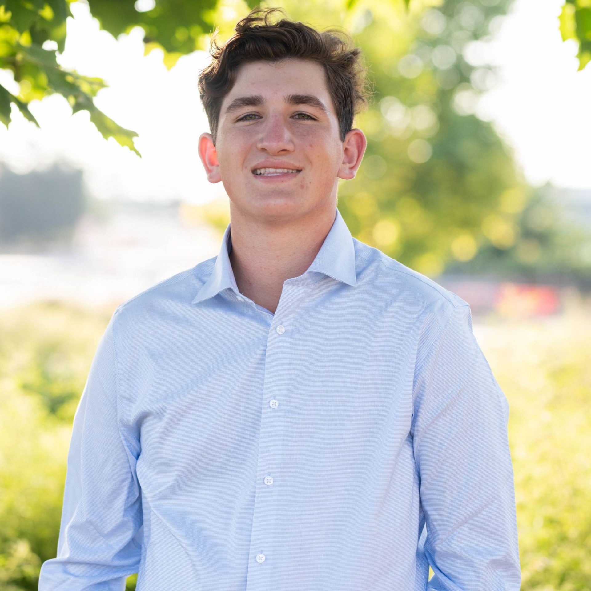Young person smiling in a light blue striped shirt outdoors with blurred greenery behind.