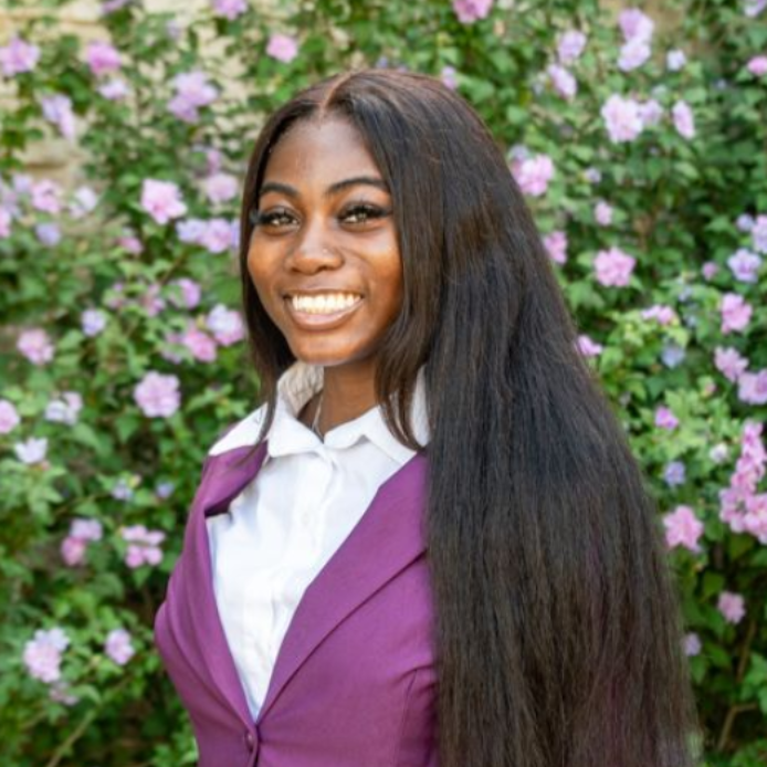 Smiling person in a purple blazer and white shirt standing before a flowering green wall