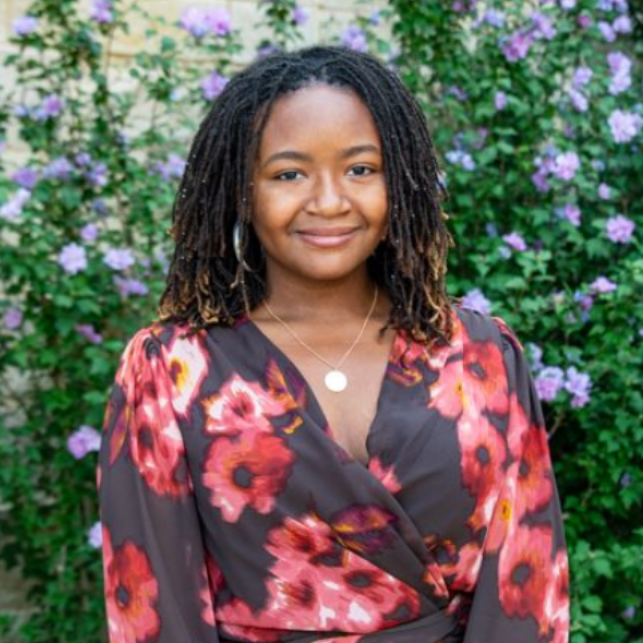 Smiling person in a floral dress standing before a leafy wall with purple flowers