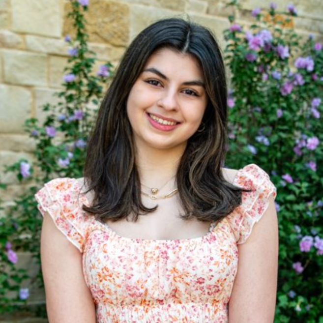 Smiling person in a pink floral dress standing outdoors by a stone wall and purple flowers