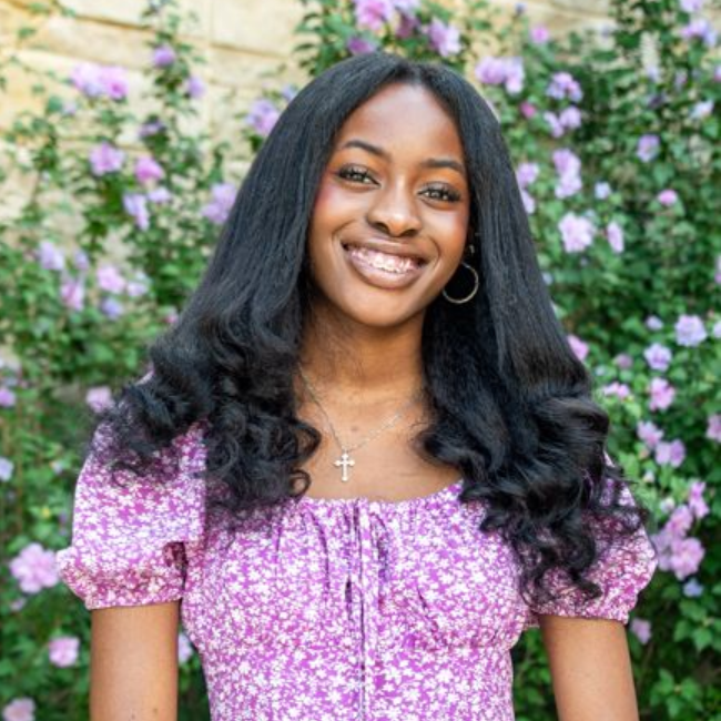 Smiling woman in a purple dress standing before a flower-covered wall