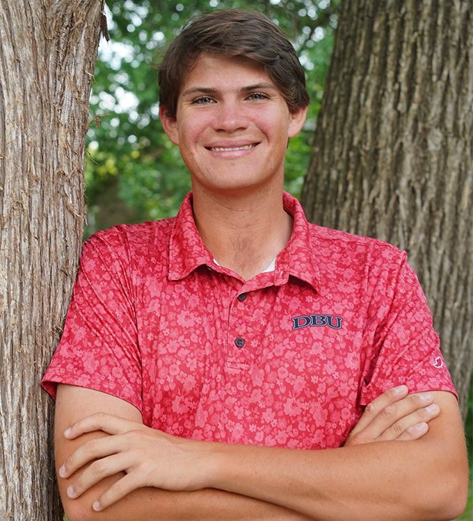 Person in a red patterned shirt standing with arms crossed between two trees, smiling.