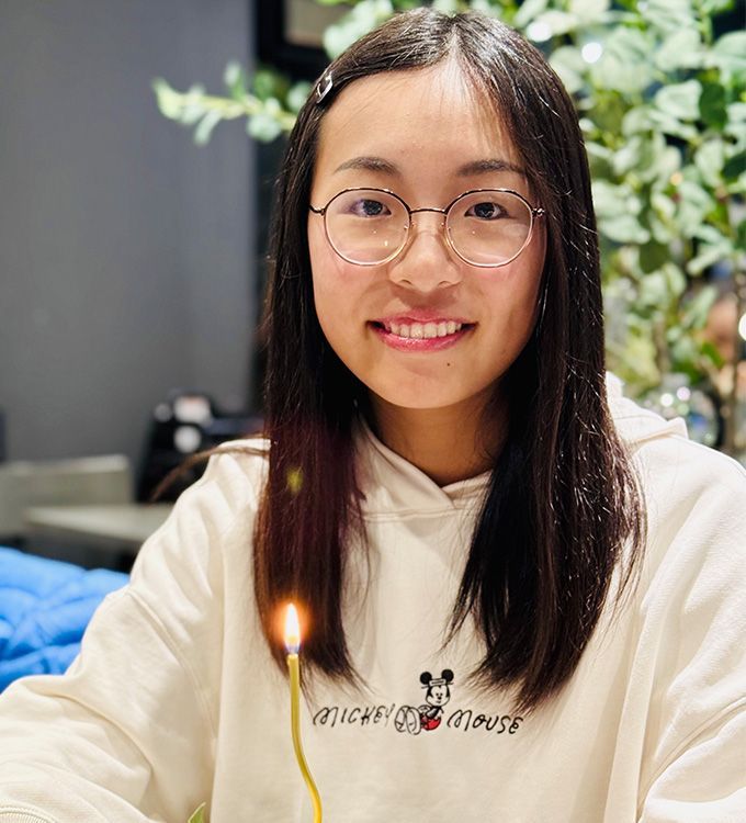 Person wearing glasses and a cream hoodie smiles near a small lit candle indoors.