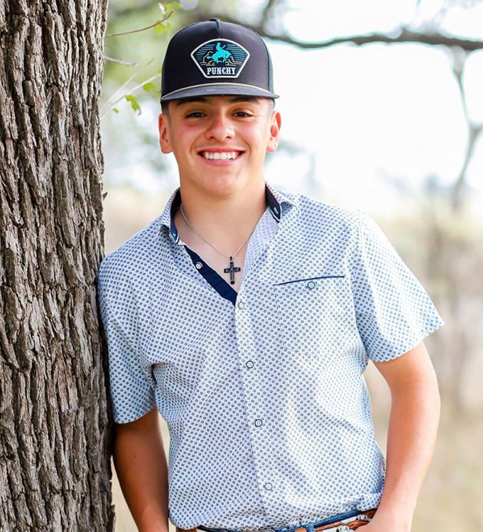 Person in a black cap and patterned shirt leans against a tree outdoors, smiling.