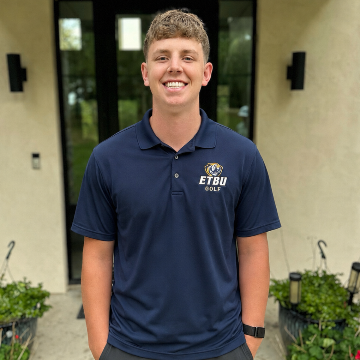Smiling person in a navy polo shirt standing outdoors in front of a building entrance.