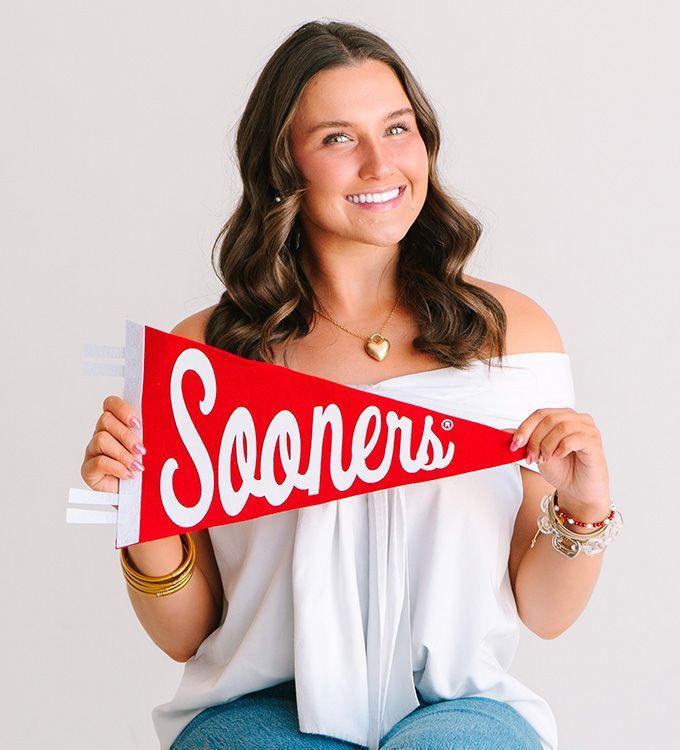 Woman holding a red Sooner's pennant and smiling in a white off-shoulder top