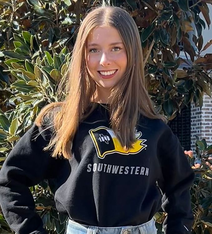 Smiling person in a black Southwestern sweatshirt standing outdoors by trees and brick wall
