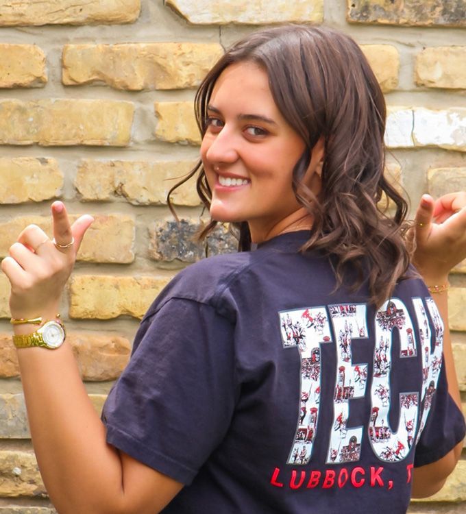 Smiling person in navy T-shirt pointing at the back, standing before a stone wall