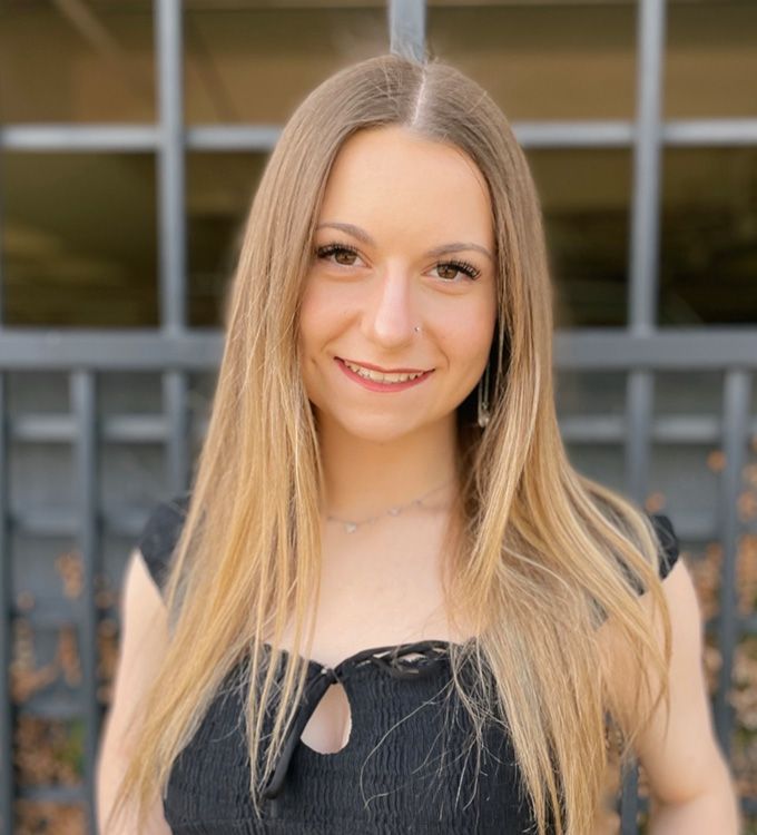 Smiling person with long blonde hair in a black top, standing outdoors before a building.