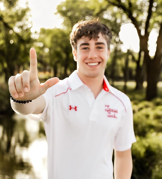 Smiling person in a white polo shirt holding up one finger outdoors in a sunny park