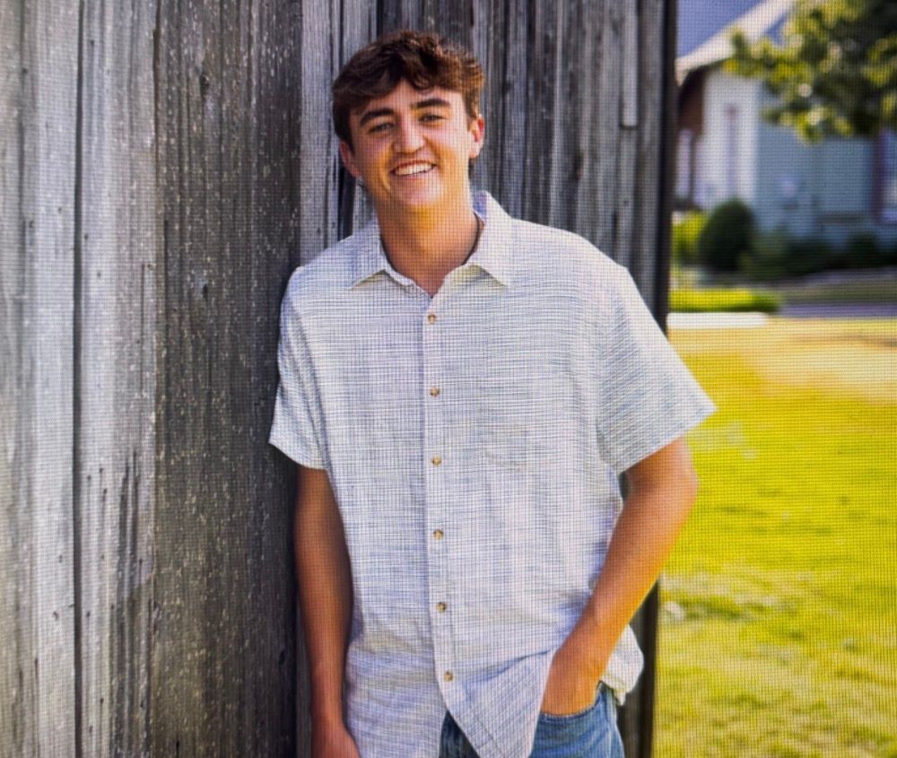 Smiling person in a light shirt leaning against a weathered wooden wall outdoors.