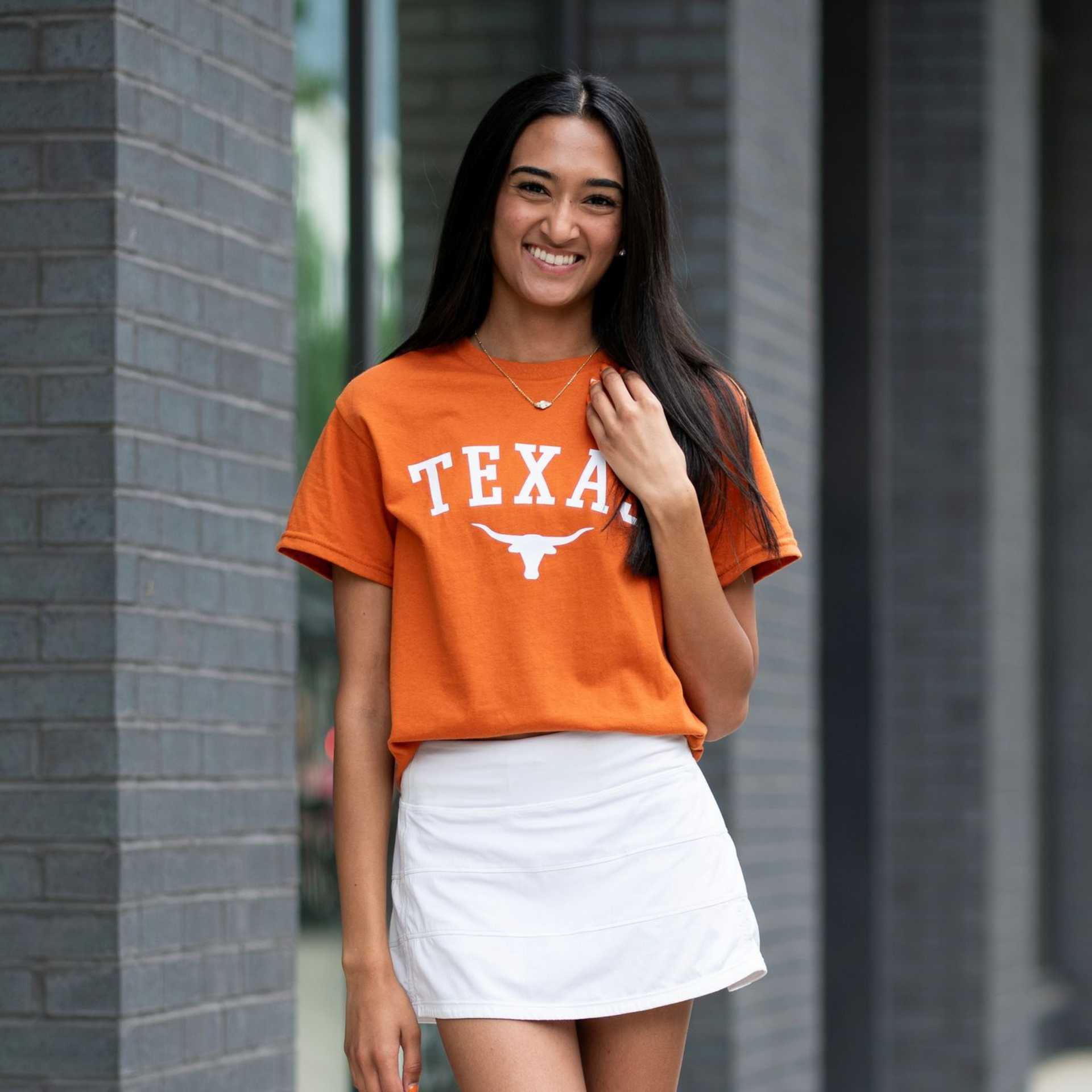 Woman in orange Texas shirt and white skirt standing outdoors by a column, smiling