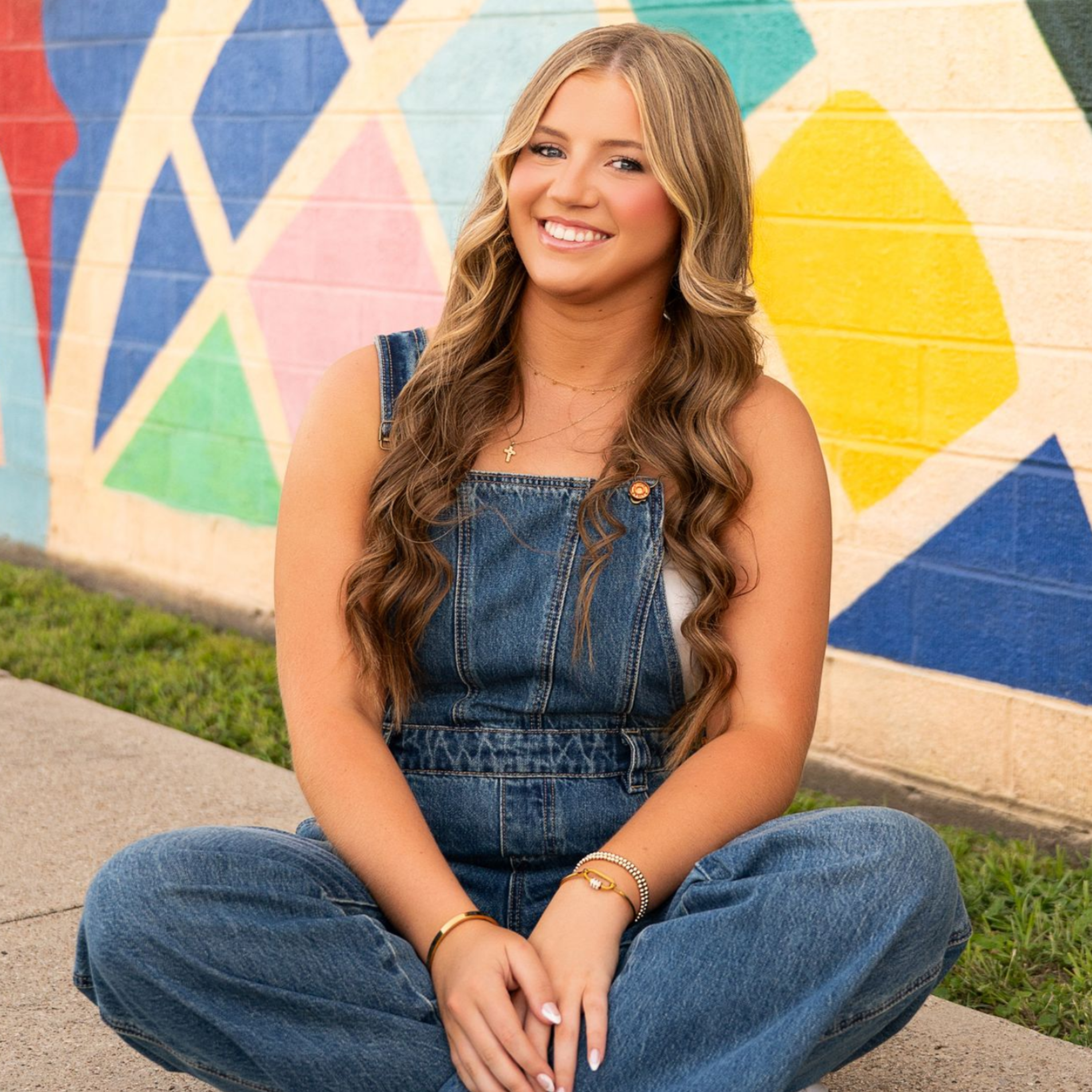 Smiling person in denim overalls sitting cross-legged before a colorful mural